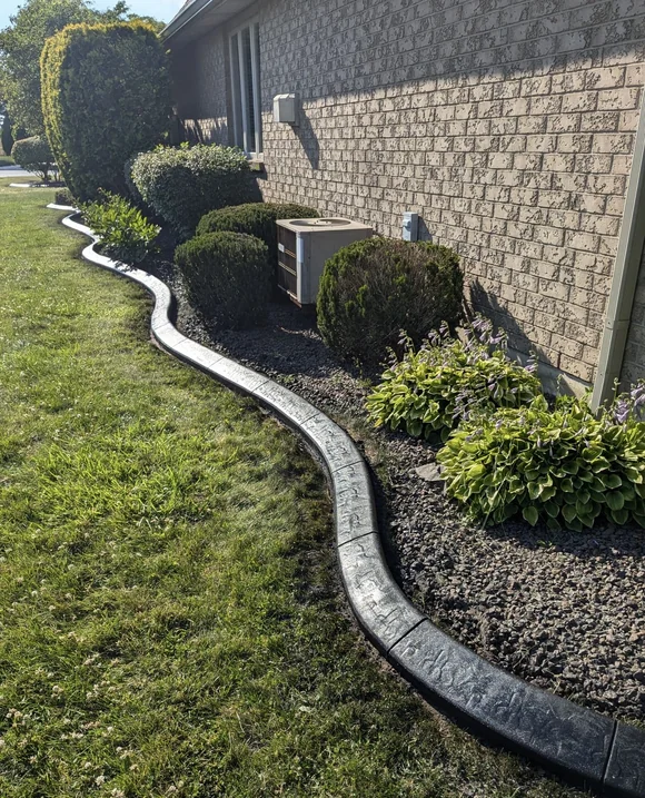 Charcoal stamped concrete curbing winding along a side garden bed with shrubs, hostas and black mulch next to a brick home in Windsor Essex County.