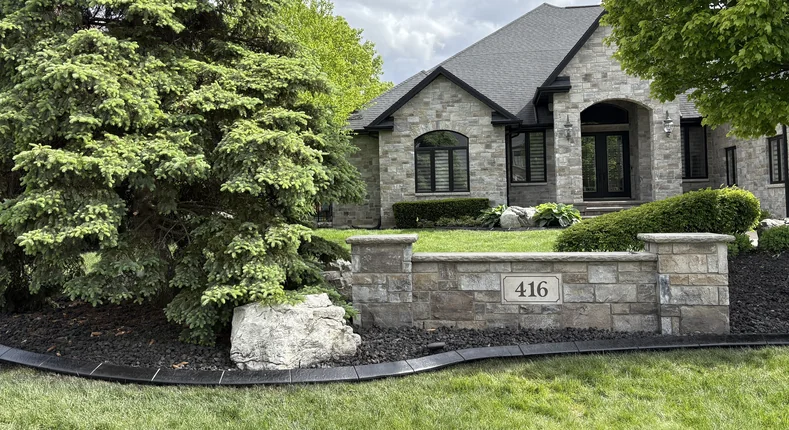 Charcoal stamped concrete curbing accenting a stone-faced luxury home with a built stone address pillar and mature spruce.