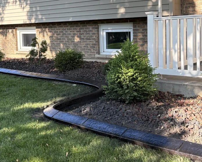 Dark brown stamped concrete curb winding along a brick house foundation with evergreen shrubs and red lava rock mulch.