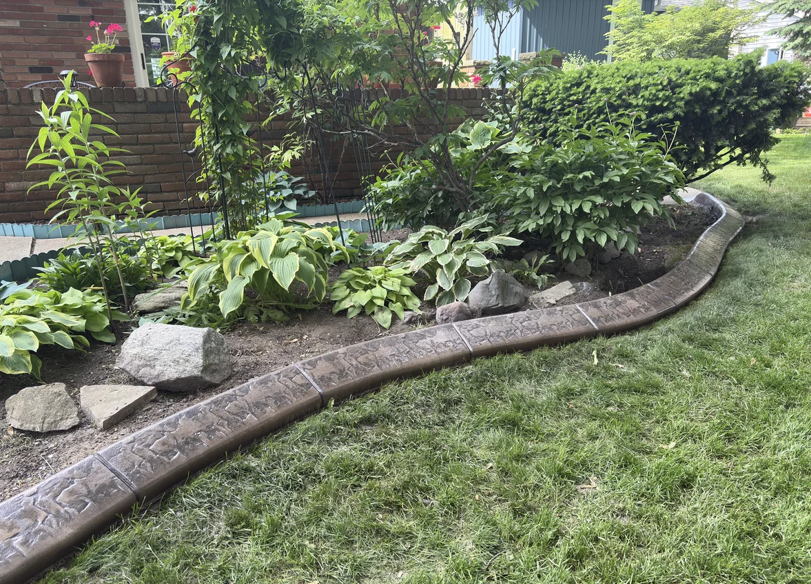 Warm brown stamped concrete curb following the edge of a perennial garden with hostas, boulders and climbing roses.