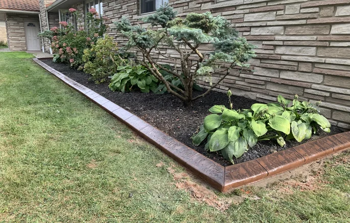Rustic brown stamped concrete curb bordering a stone-veneer home with a conifer, hydrangea and hostas along the foundation.